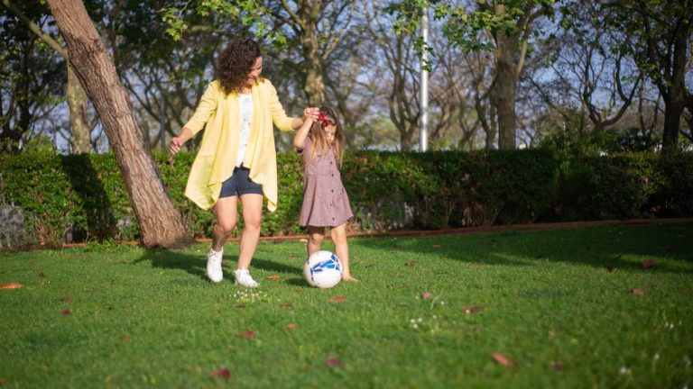 A mother and daughter enjoying a sunny day playing soccer in a lush green park.