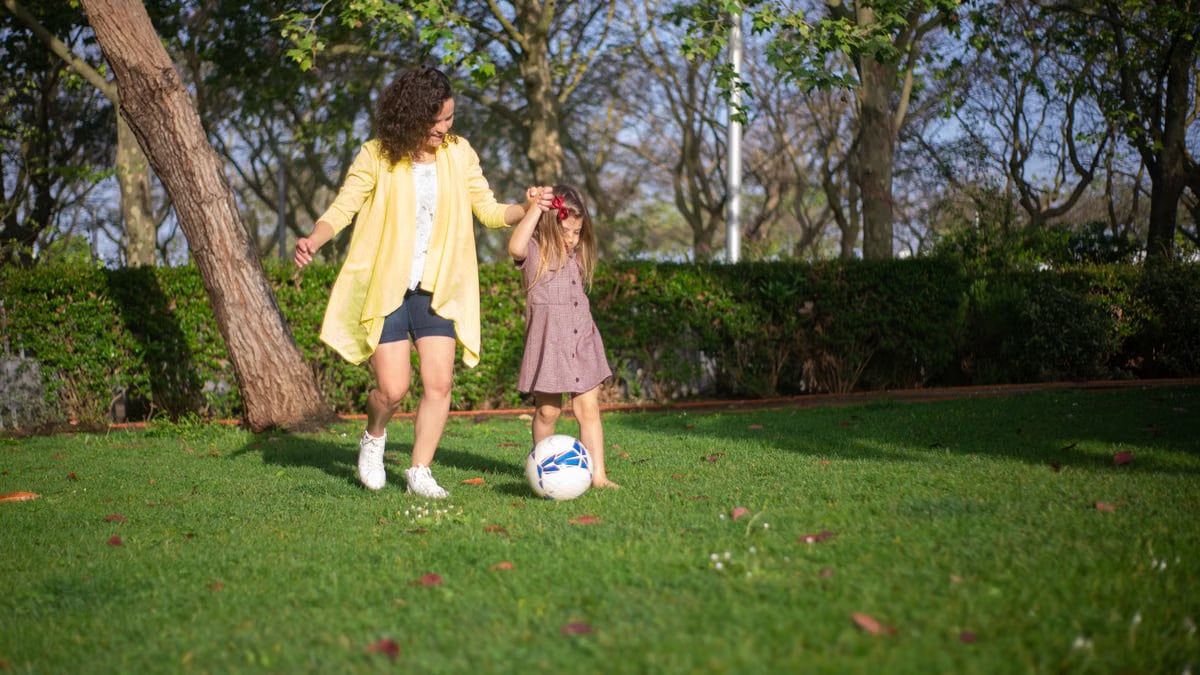 A mother and daughter enjoying a sunny day playing soccer in a lush green park.